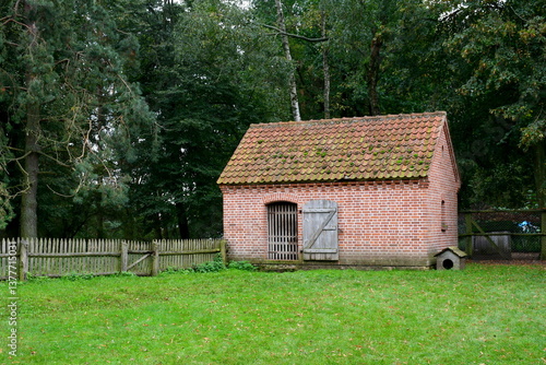 A view of a small hut, shack, shelter or barn made out of red brick and having an angled, tiled roof located next to a vast lawn, a wooden fence and a small wooden dog house seen in Poland