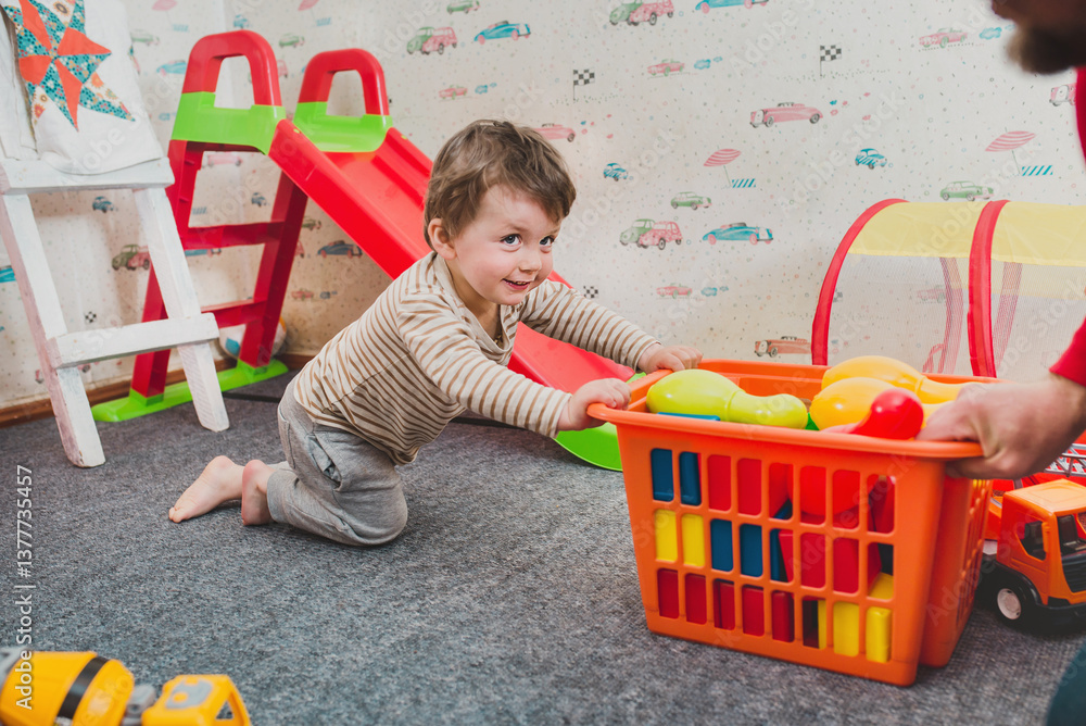 Naklejka premium Little boy 2 years old playing in the nursery at home. Shaking plastic toys out of the basket. Mess from a small child. Fun educational games. Independent play