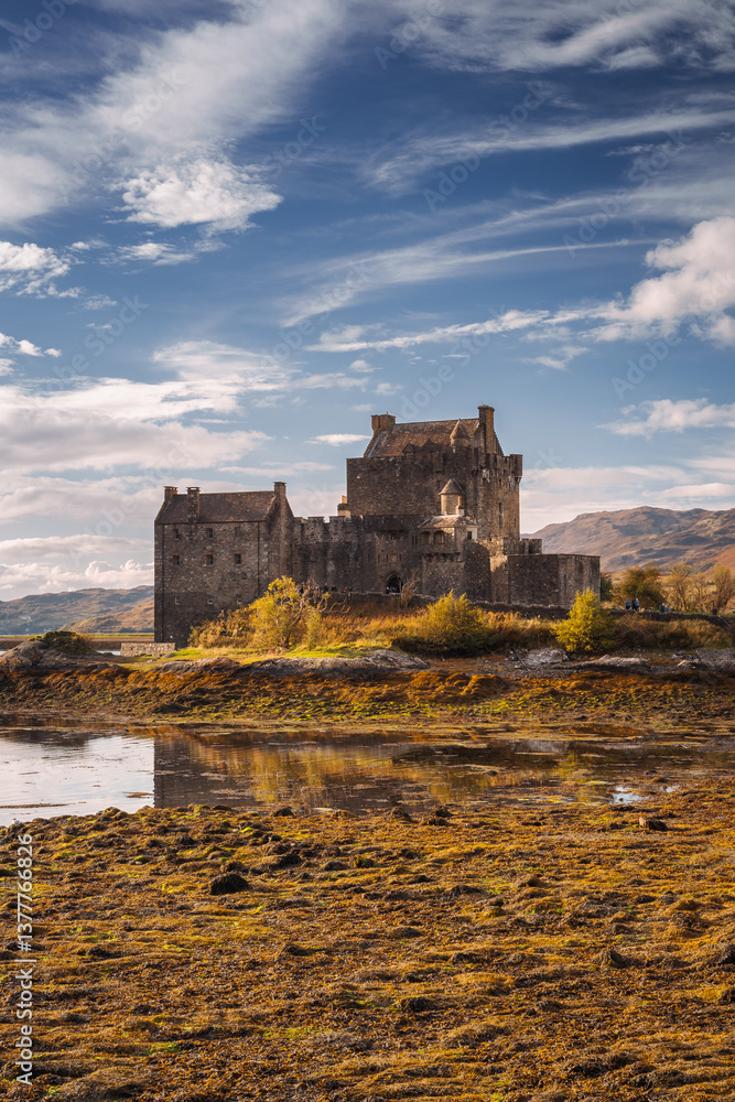 Eilean Donan Castle stands majestically amid autumn hues, its reflection shimmering in calm waters. Misty Highlands and golden trees create a scene of timeless beauty.