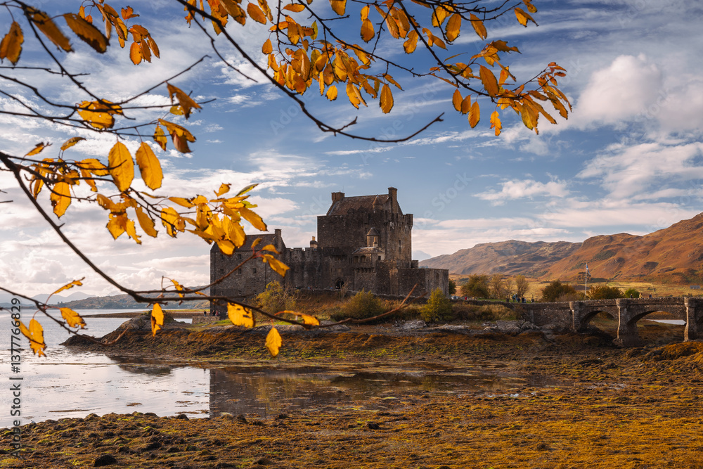 Eilean Donan Castle stands majestically amid autumn hues, its reflection shimmering in calm waters. Misty Highlands and golden trees create a scene of timeless beauty.