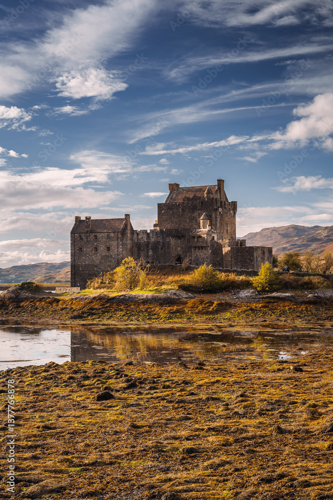 Eilean Donan Castle stands majestically amid autumn hues, its reflection shimmering in calm waters. Misty Highlands and golden trees create a scene of timeless beauty.