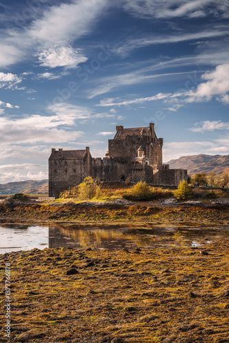 Obraz na płótnie Eilean Donan Castle stands majestically amid autumn hues, its reflection shimmering in calm waters. Misty Highlands and golden trees create a scene of timeless beauty. w salonie