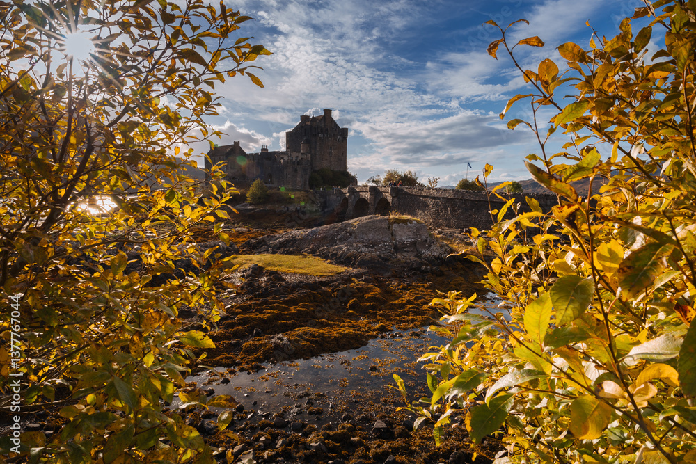 Eilean Donan Castle stands majestically amid autumn hues, its reflection shimmering in calm waters. Misty Highlands and golden trees create a scene of timeless beauty.