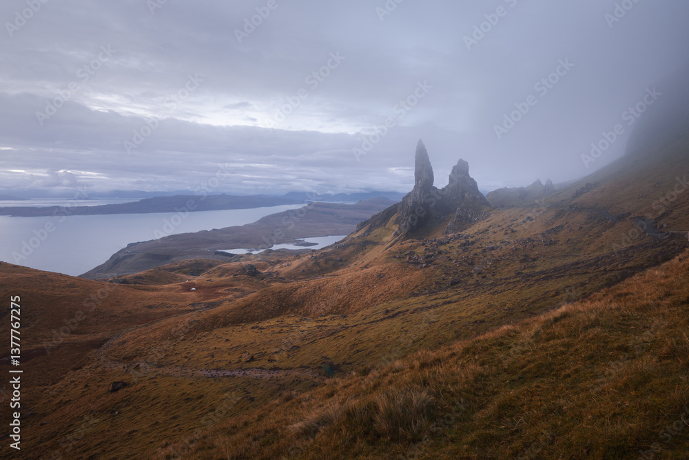 The Old Man of Storr emerges from autumn mist, its towering pinnacles glowing in golden morning light. A mystical, otherworldly Highland scene.
