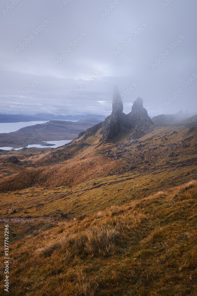 The Old Man of Storr emerges from autumn mist, its towering pinnacles glowing in golden morning light. A mystical, otherworldly Highland scene.