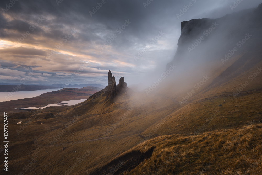 The Old Man of Storr emerges from autumn mist, its towering pinnacles glowing in golden morning light. A mystical, otherworldly Highland scene.