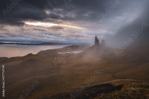 The Old Man of Storr emerges from autumn mist, its towering pinnacles glowing in golden morning light. A mystical, otherworldly Highland scene.