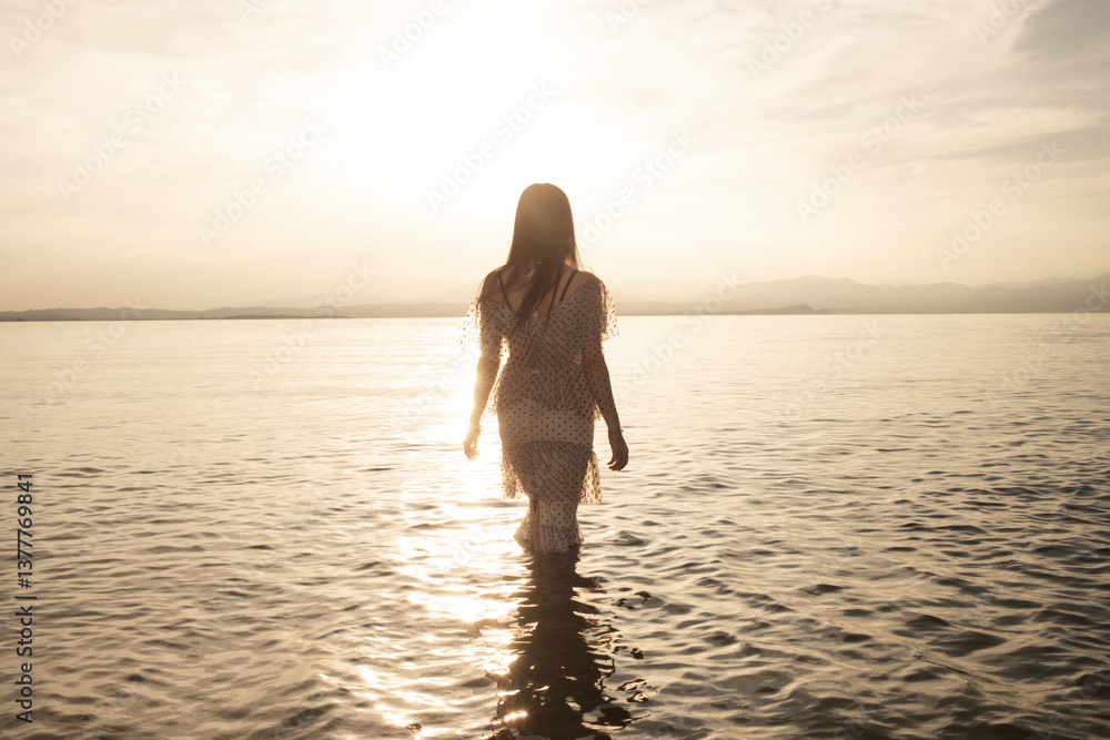 Elegant woman walking among the waves at sunset, illuminated by the golden light of the sun Elegant woman walking among the waves at sunset, illuminated by the golden light of the sun