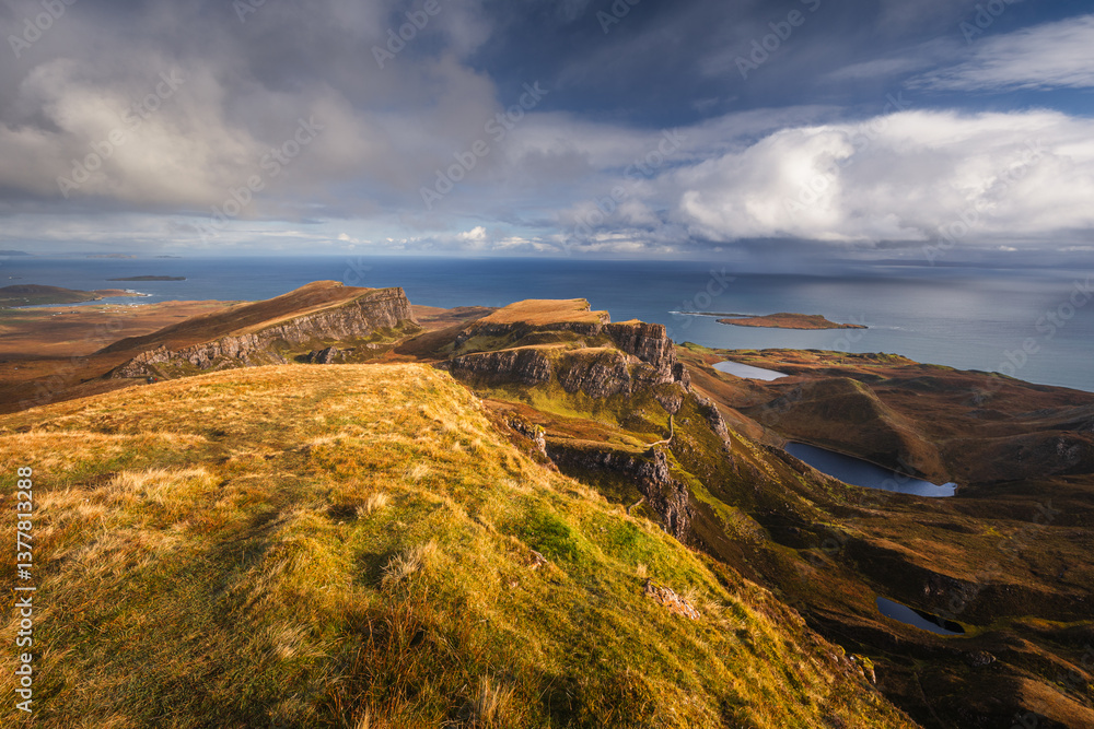 Autumn light bathes the Quiraing on Skye, casting long shadows over its dramatic cliffs and rolling hills. The landscape glows with golden hues, a breathtaking Highland scene.