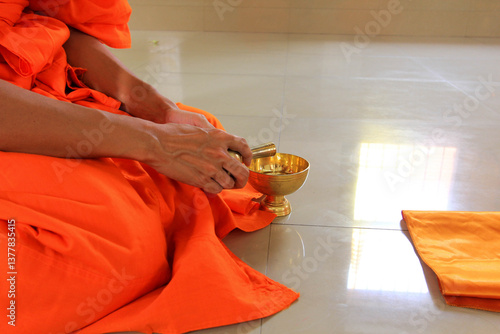 A Buddhist monk in vibrant orange robes performs a ritual, pouring water from a golden vessel into a small bowl. The solemn act is captured in a serene setting.