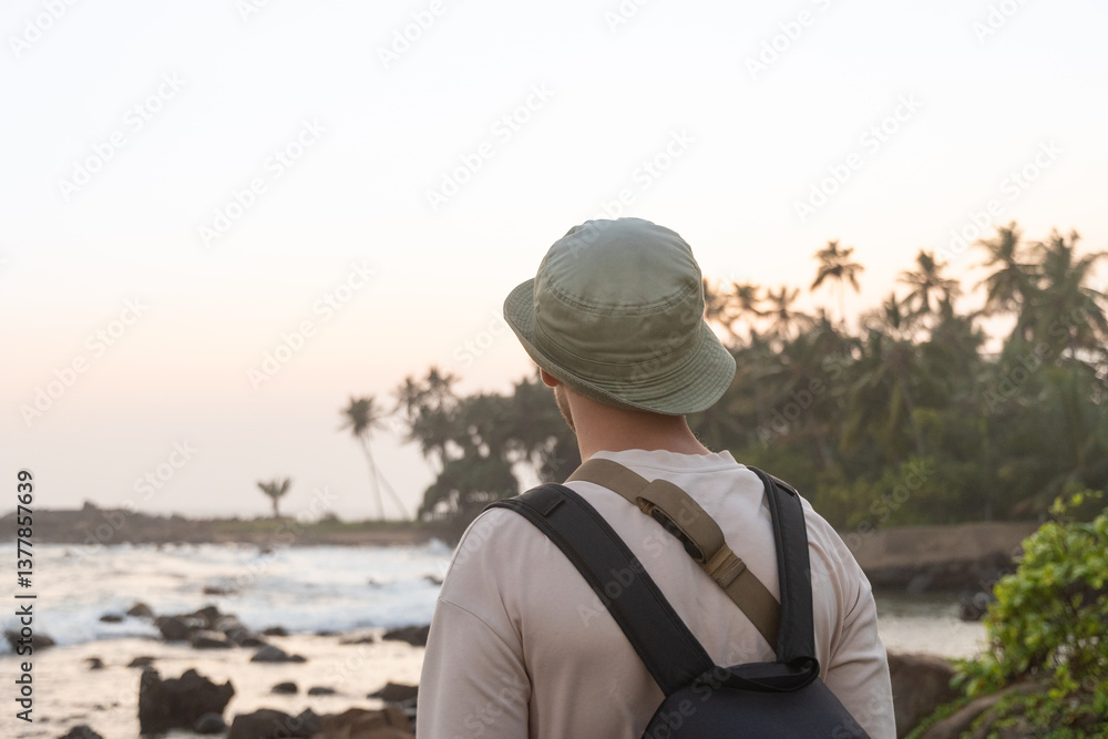 Scenic view of traveler wearing hat and backpack, gazing at ocean during sunset. Palm trees, rocky shore, ocean and serene atmosphere. Travel summer vacation.