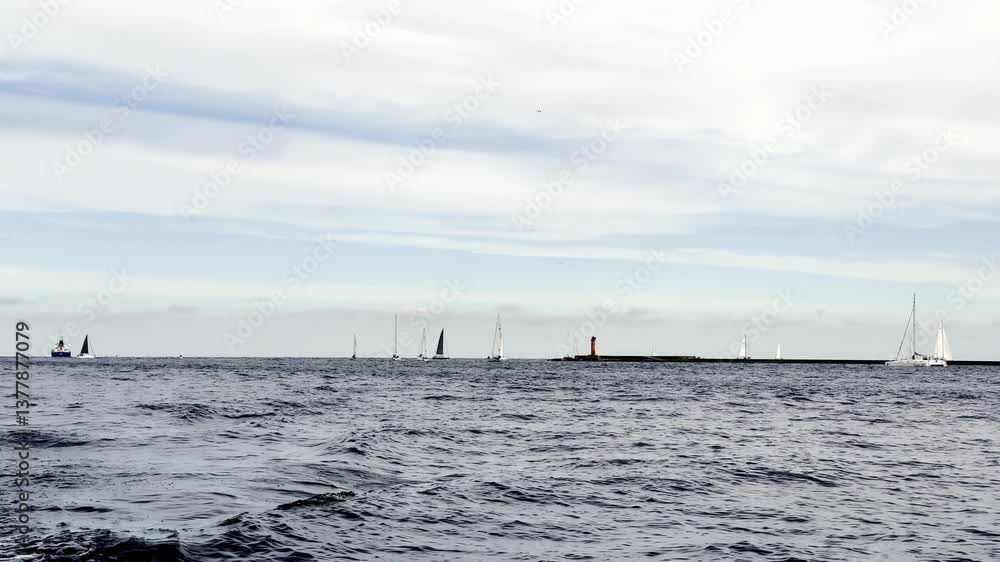 A wide view of sailboats gliding across the calm surface of the Baltic Sea near a distant lighthouse. The sky above is softly clouded, adding to the tranquil maritime atmosphere. A wide view of sailboats gliding across the calm surface of the Baltic Sea near a distant lighthouse. The sky above is softly clouded, adding to the tranquil maritime atmosphere.
