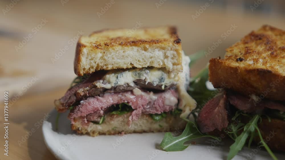 A sandwich with meat and greens is cut in half and placed on a white plate. The sandwich is served on a wooden table A sandwich with meat and greens is cut in half and placed on a white plate. The sandwich is served on a wooden table