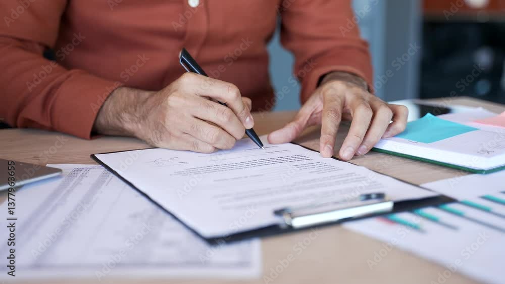 Close up of a male's hand signing documents at a desk at a workplace in a business office. The boss or executive looks through the folder with documentation and signs the contract with a pen