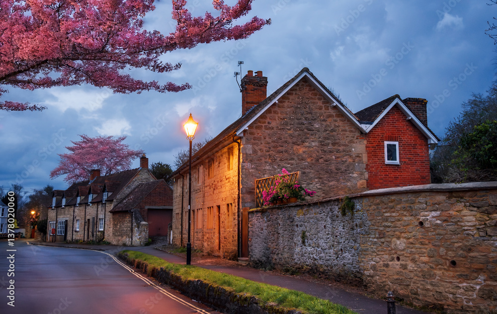 A charming evening in Iffley, Oxford, with historic Cotswold stone cottages, glowing streetlights, and vibrant cherry blossoms in spring season, UK