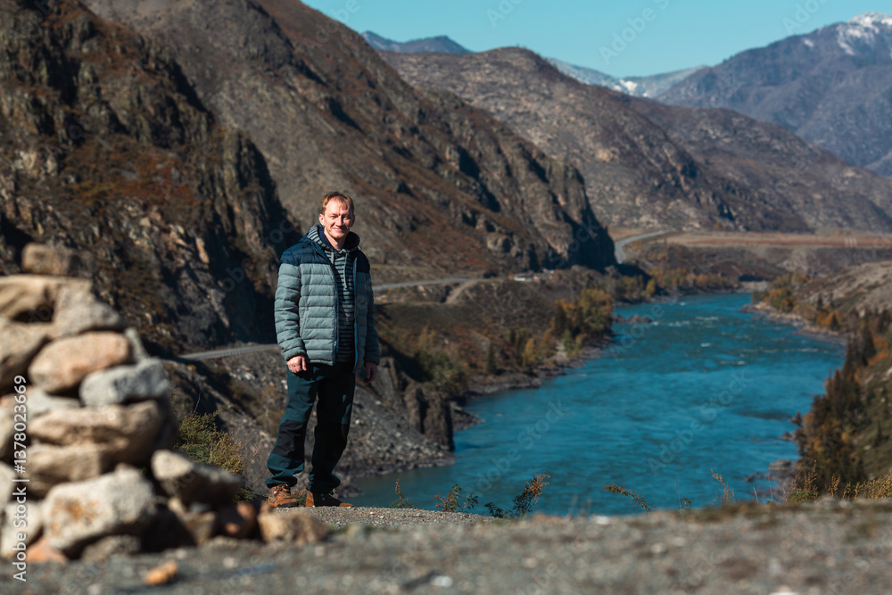 A man stands by a river in the mountains of the Altai Republic, Russia, surrounded by a rugged landscape.