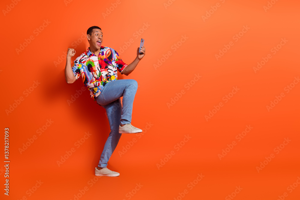 Energetic young man celebrating success with smartphone in hand, wearing colorful shirt against vibrant orange background Energetic young man celebrating success with smartphone in hand, wearing colorful shirt against vibrant orange background