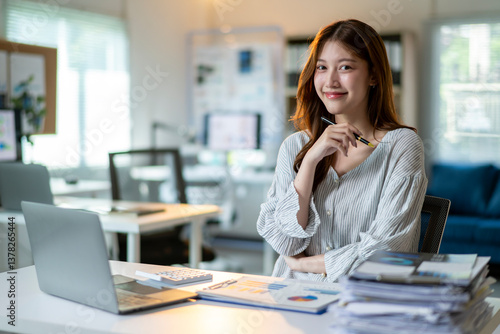 A woman is sitting at a desk with a laptop and a stack of papers