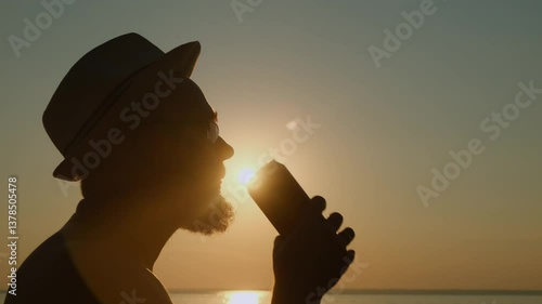 Adult bearded man in a hat drinking beer from a can by the sea at sunset. A refreshing sip of cold drink on a hot summer evening on vacation.