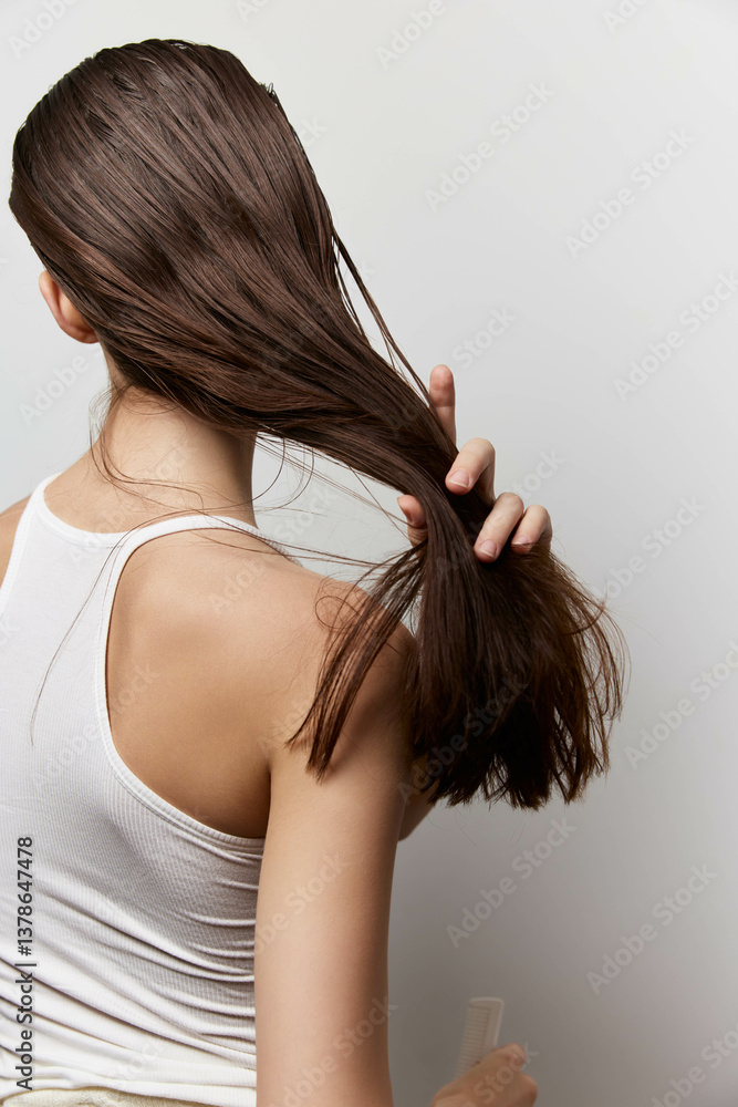 Naklejka premium Woman with long brown hair, gently holding it while facing a neutral background, showcasing smooth strands and natural shine Ideal for beauty and haircare concepts