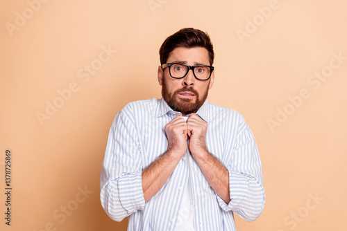 Worried young man with glasses clutching shirt, expressing nervousness against a neutral beige background