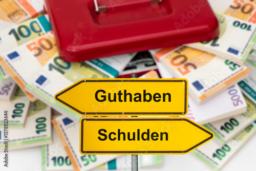 Vor einer Geldkassette mit vielen Bündel Euro Banknoten steht ein Wegweiser beschriftet mit Guthaben und Schulden - FOTOMONTAGE