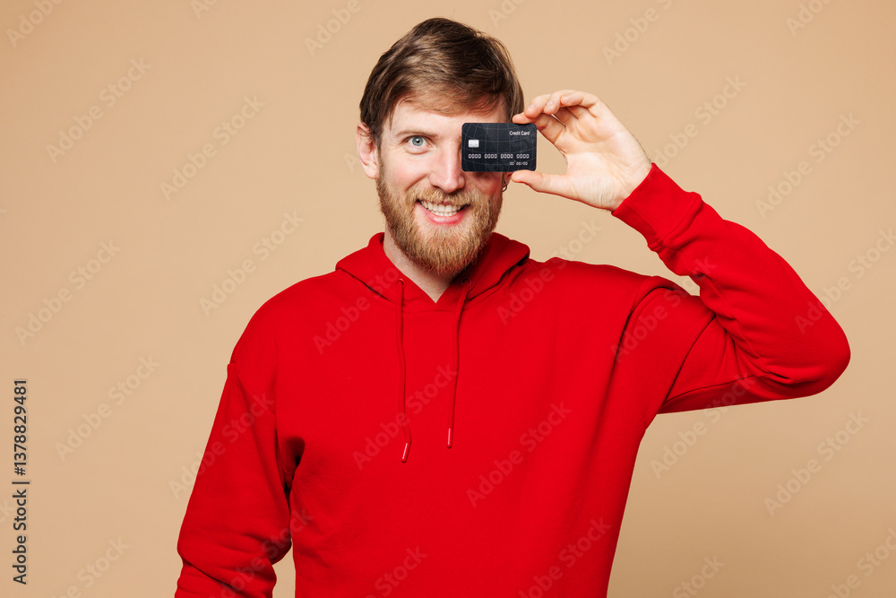 Young Caucasian man he wear red hoodie casual clothes hold in hand cover eye face with mock up of credit bank card isolated on pastel plain light beige background studio portrait. Lifestyle concept. Young Caucasian man he wear red hoodie casual clothes hold in hand cover eye face with mock up of credit bank card isolated on pastel plain light beige background studio portrait. Lifestyle concept.