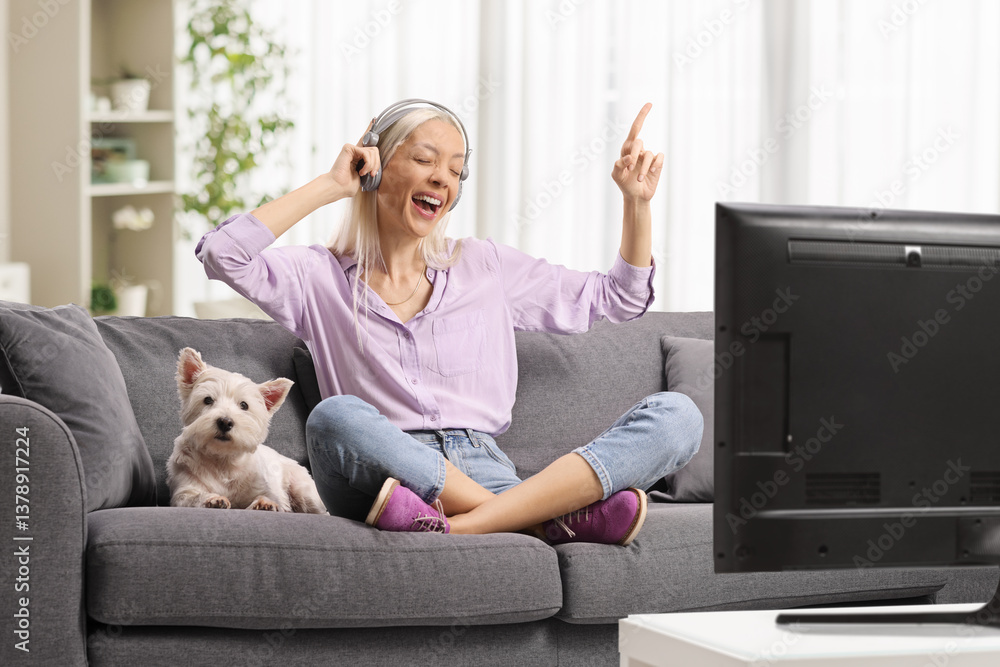 Happy woman sitting on a sofa with a westie terrier dog and listening to music in front of tv Happy woman sitting on a sofa with a westie terrier dog and listening to music in front of tv