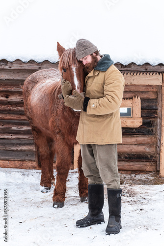A man with a beard stands hugging a horse and looks at it, winter, snow, village, peasant, farm, wooden house.