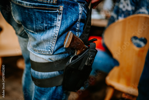 Photos Close-up of a person in blue jeans wearing a leg holster with a handgun, likely as part of a cowboy or western-themed costume at an indoor event