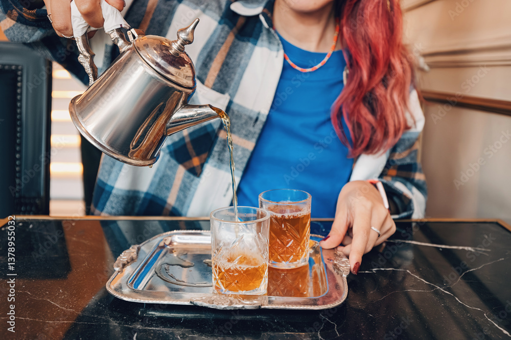 Woman pouring hot tea from a metal teapot into two crystal glasses placed on an elegant silver tray