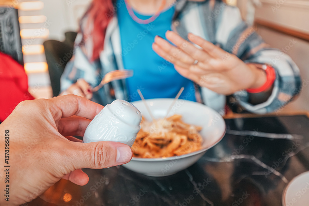 Hand holding salt shaker near plate of pasta and woman making refuse no gesture with hand in restaurant