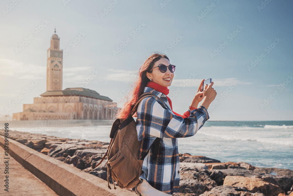 Smiling tourist capturing the stunning Hassan II Mosque by phone at sunset, surrounded by the vibrant atmosphere of Casablanca, Morocco