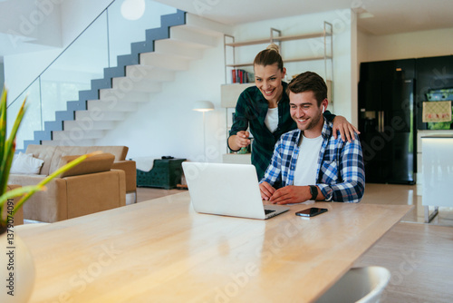 A young married couple is talking to parents, family and friends on a video call via a laptop while sitting in the living room of their modern house.