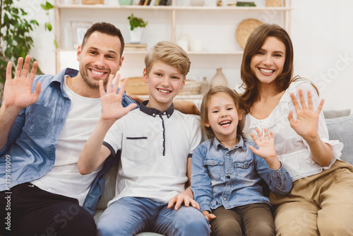 Hello. Happy Family Waving Hand Sitting Together On Couch At Home, Smiling To Camera