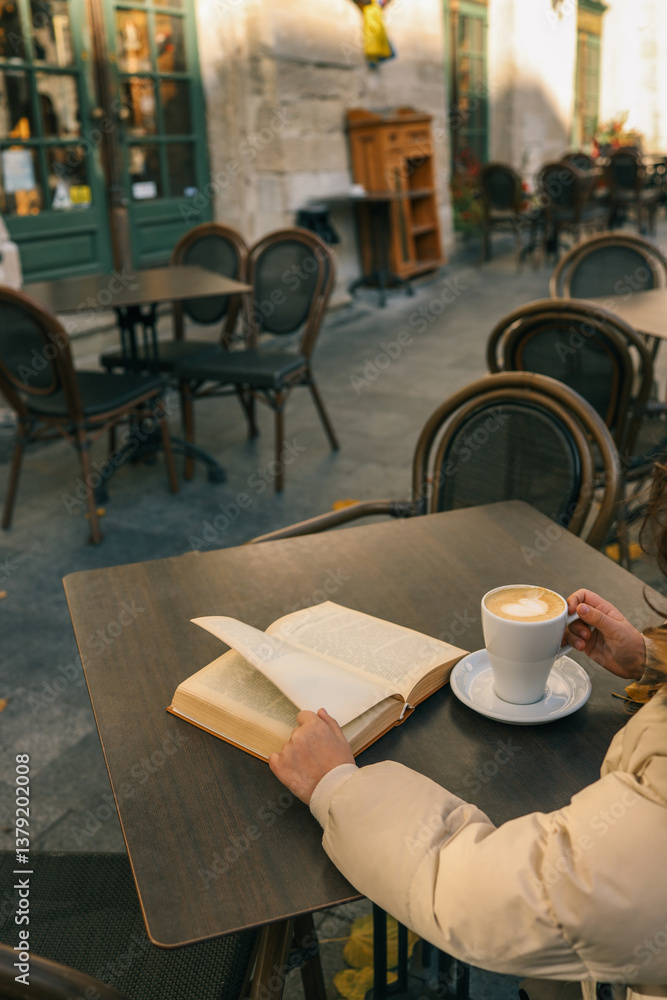A person enjoys a latte and a book at an outdoor café table on a quiet street. Cozy autumn or winter vibe with warm light and European-style architecture in the background