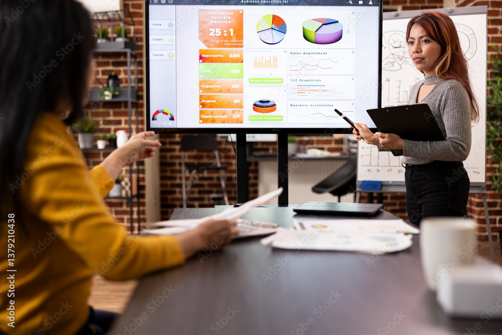 Female colleagues collaborating together on preparing marketing strategies in brick wall office. Asian business advisor stands near digital monitor, listening to coworker give insights during meeting. Female colleagues collaborating together on preparing marketing strategies in brick wall office. Asian business advisor stands near digital monitor, listening to coworker give insights during meeting.
