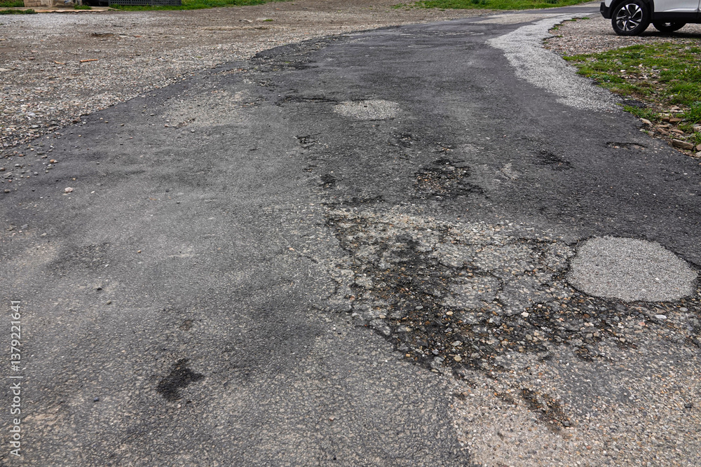 A cracked and uneven road stretches through a suburban landscape, displaying various potholes and patches.