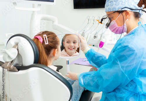 Pediatric dentist showing mirror to smiling girl in dental clinic