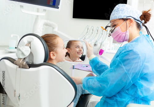 Pediatric dentist showing mirror to smiling girl in dental clinic