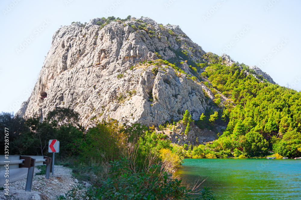 River Cetina and mountain in Omis from Croatia. Majestic rocky mountain adorned with greenery, set against a serene lake with clear sky above, creating a harmonious and tranquil natural landscape