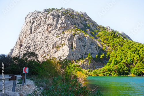 River Cetina and mountain in Omis from Croatia. Majestic rocky mountain adorned with greenery, set against a serene lake with clear sky above, creating a harmonious and tranquil natural landscape