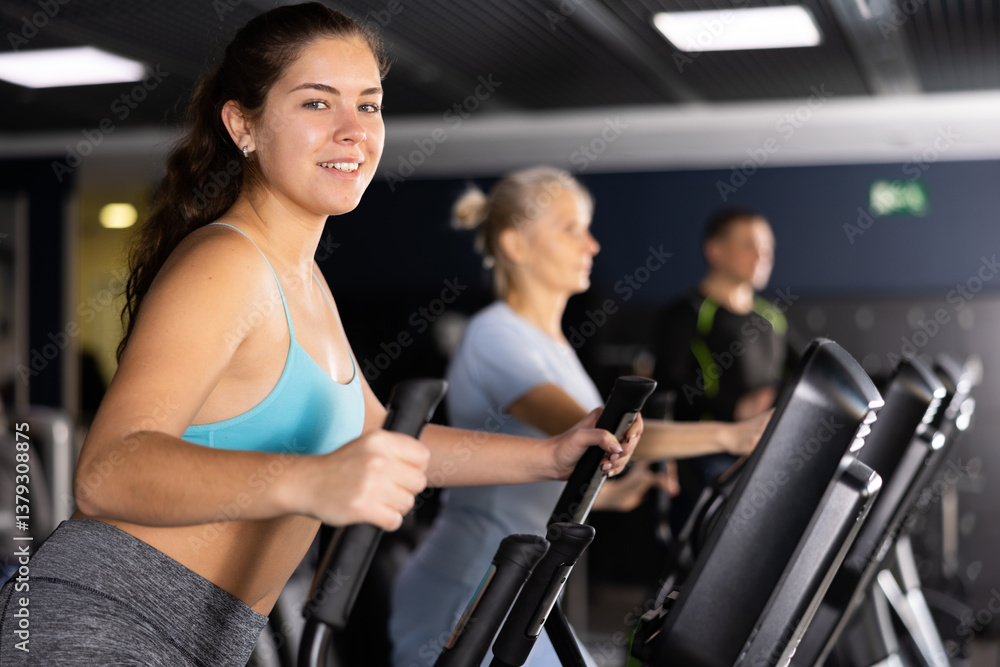 Focused young girl warming up on elliptical machine before strength training in modern gym.. Focused young girl warming up on elliptical machine before strength training in modern gym..