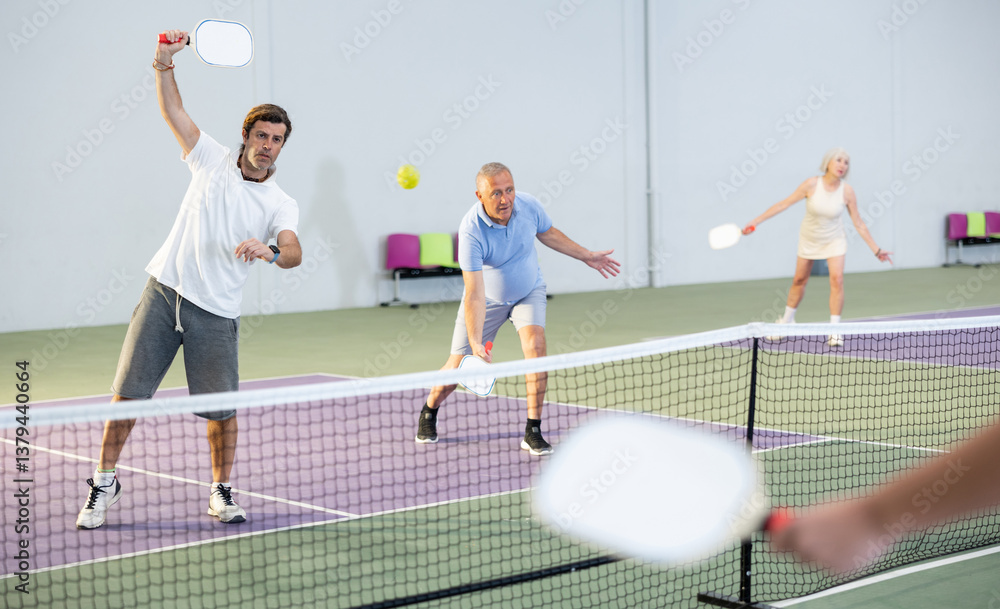 Obraz premium Portrait of concentrated adult pickleball player preparing to strike and return ball to opponents field during doubles match in team with older partner on indoor court