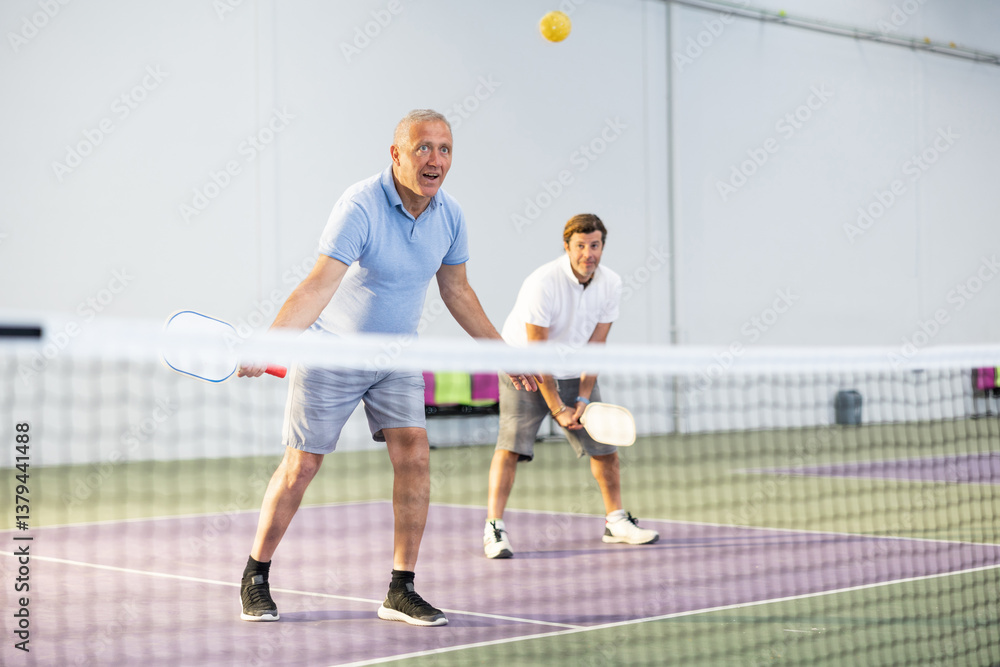 Obraz premium Portrait of sporty senior man playing doubles pickleball with male partner on indoor court, ready to hit ball. Sport and active lifestyle concept..