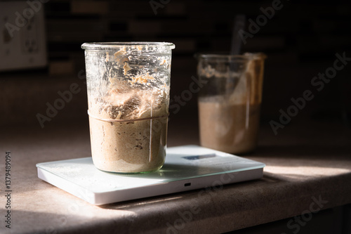 Glass container of sourdough starter on a kitchen scale with discard in the background 