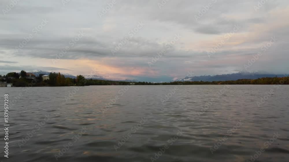 Fast Drone Glide at Low Altitude Over the Waters of Wasilla Lake, Alaska, with Orange Sky, Clouds, and Snow-Capped Mountains on the Horizon