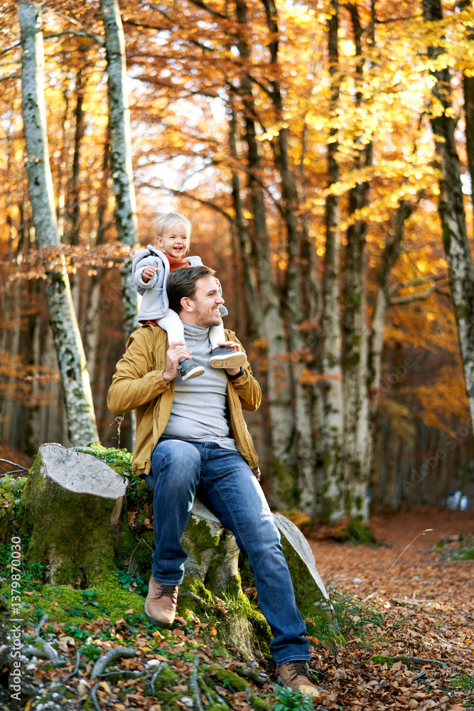 Little smiling girl sits on the shoulders of her dad sitting on a stump in an autumn park Little smiling girl sits on the shoulders of her dad sitting on a stump in an autumn park