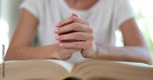 Worried woman prays holding hands over open thick old book in room. Lady with troubles seeks help in spiritual practices at home. Religion ritual