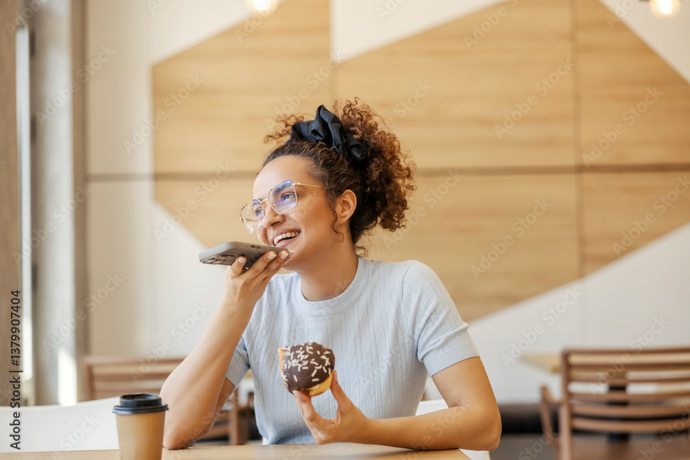 Portrait of young woman sitting in fast food restaurant with doughnut in hands and sending voice messages.